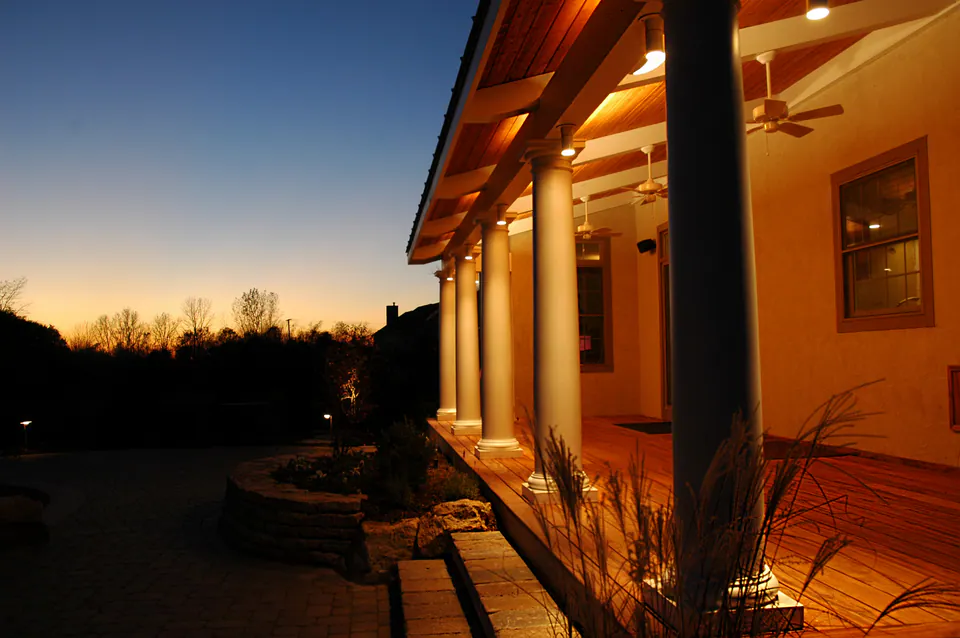 Covered porch with columns glowing at dusk beside a low stone wall and ornamental grasses